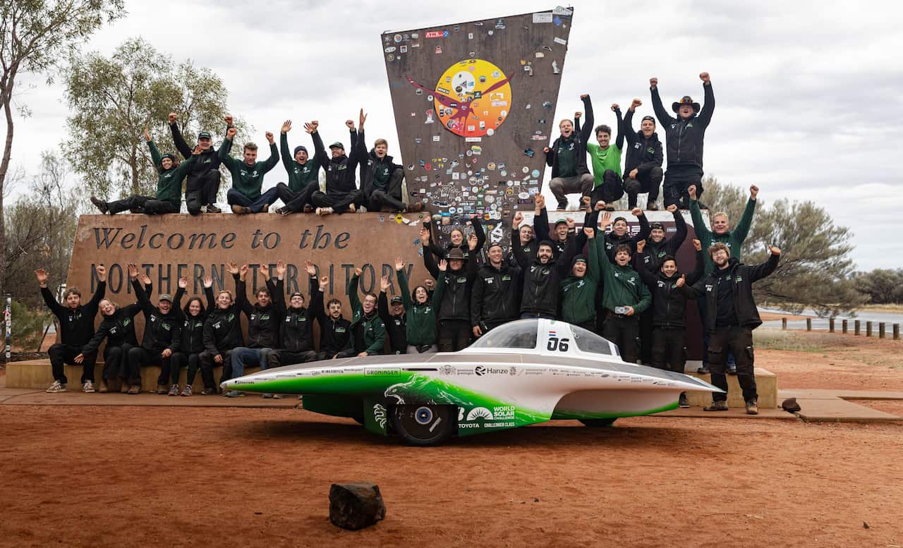 About two dozen people with their hands in the air standing in front of a sign that reads "Welcome to the Northern Territory," as well as a solar vehicle ppositioned in front of them. 