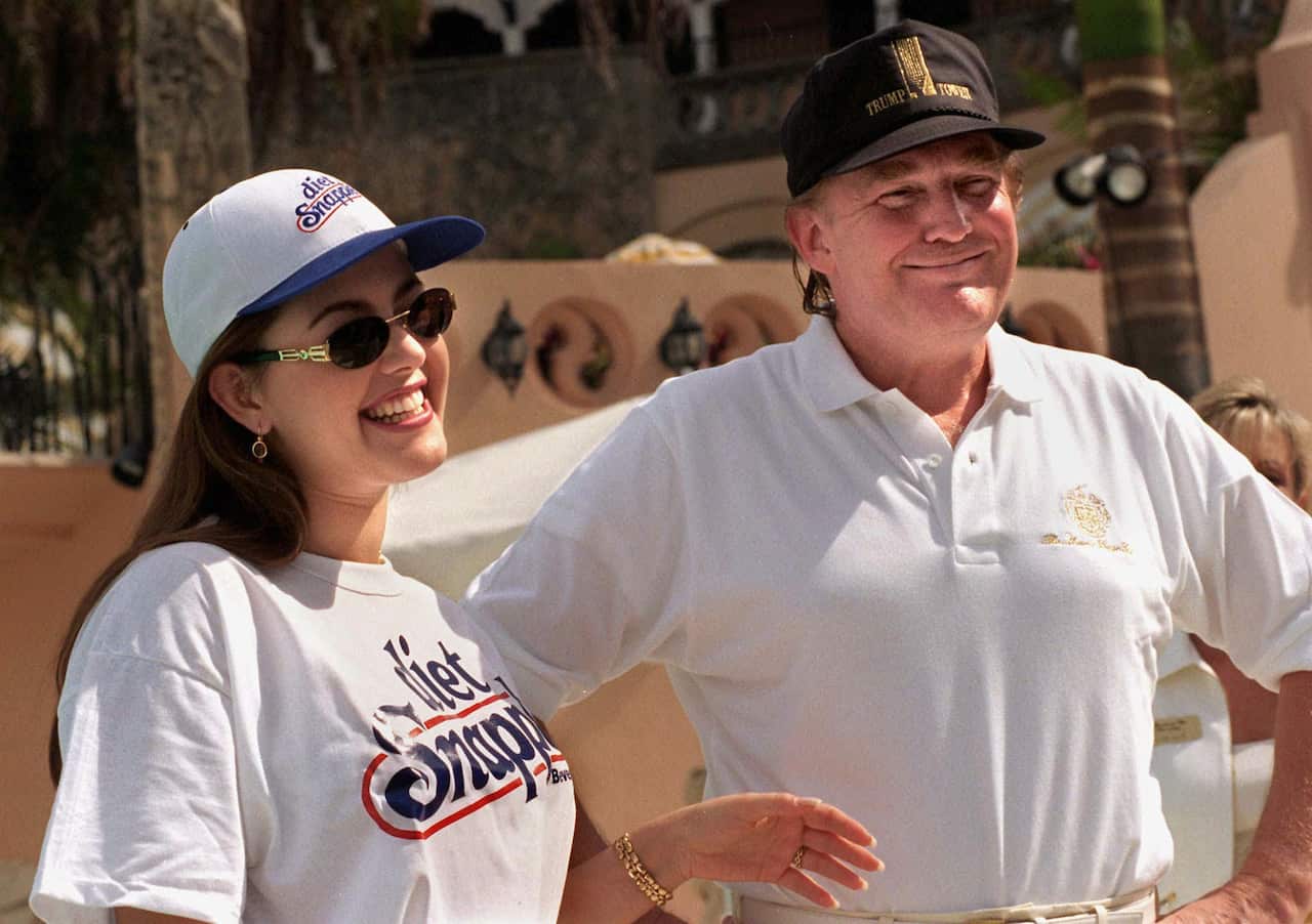 A young woman in a t-shirt, sunglasses and baseball cap stands next to a smiling, younger Donald Trump, who is also wearing a baseball hat.