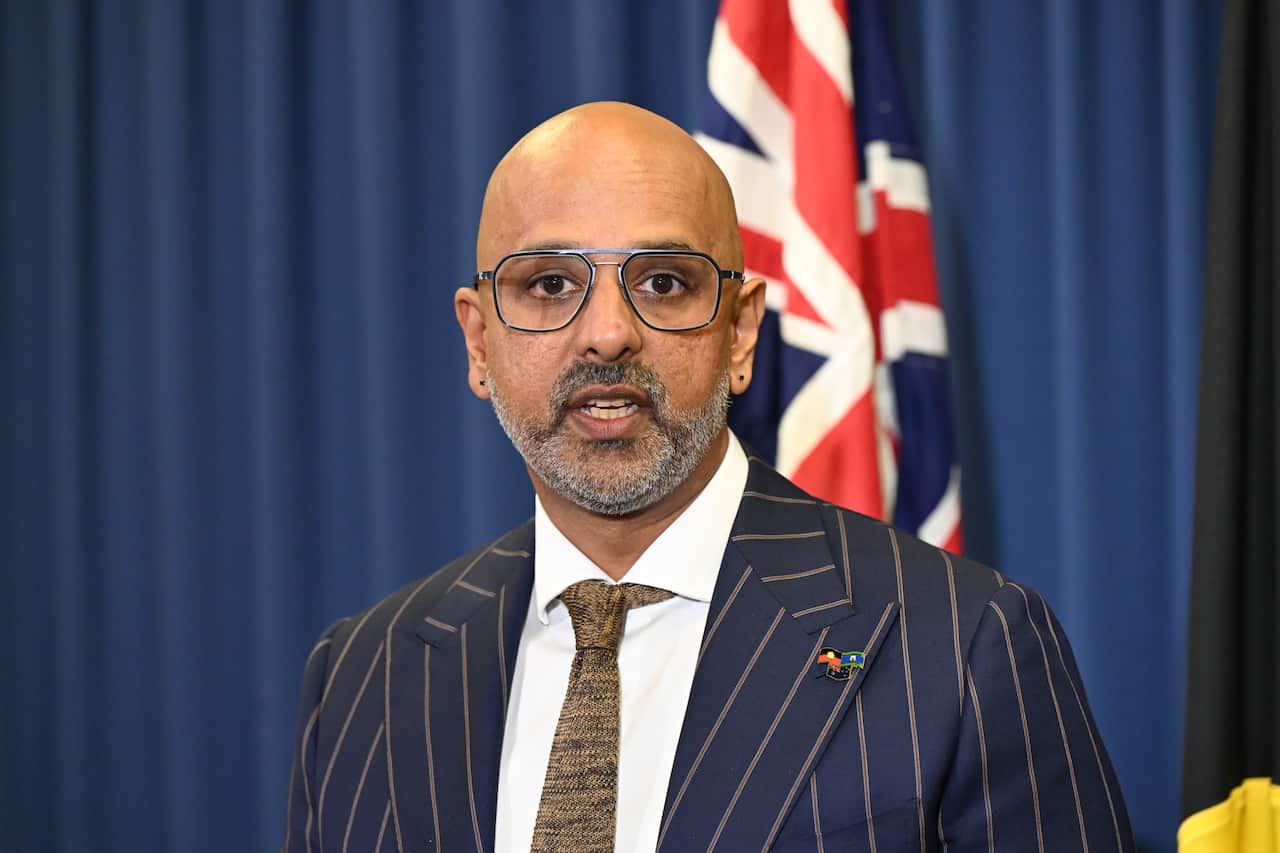 A middle-aged South Asian man wearing a pin-striped suit. An Australian flag is behind him.