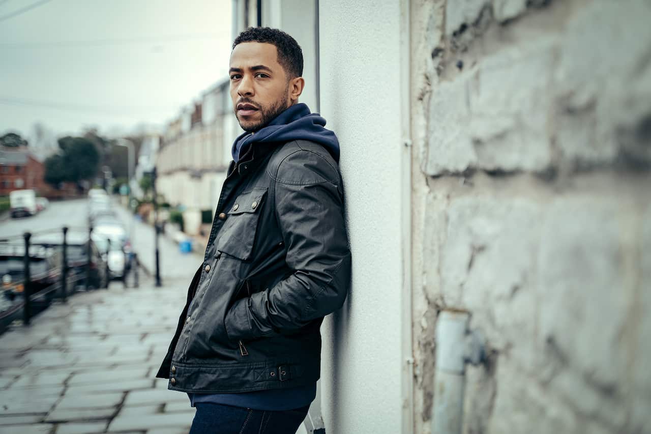 A man in blue hoodie under a dark jacket leans against a pale stone wall, looking sideways at the camera. A street stretches away behind him. 