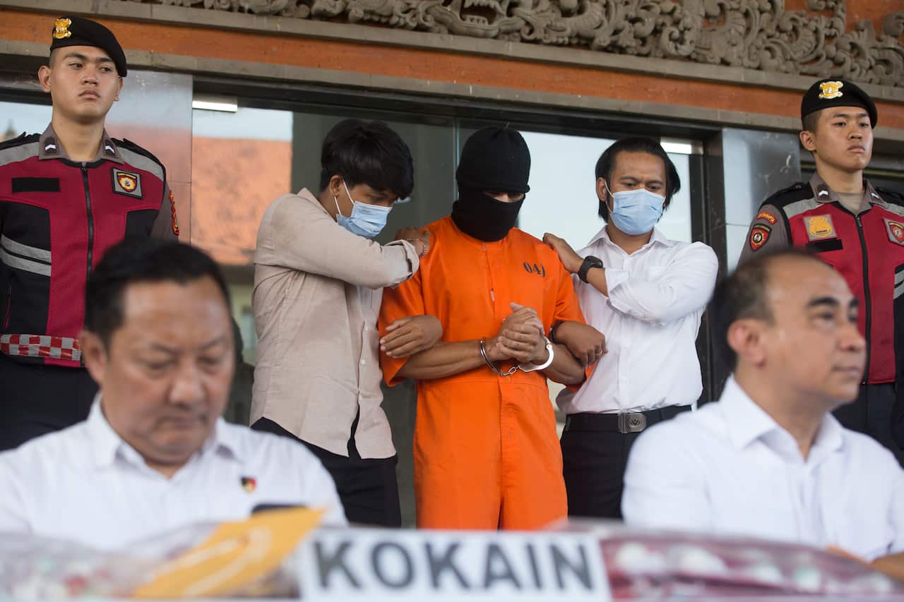 A man in an orange jumpsuit and black balaclava stands between two men who are holding him. His hands are handcuffed in front of him. There are two policemen in dark red jackets and berets either side of them. Two other men sit in the foreground.