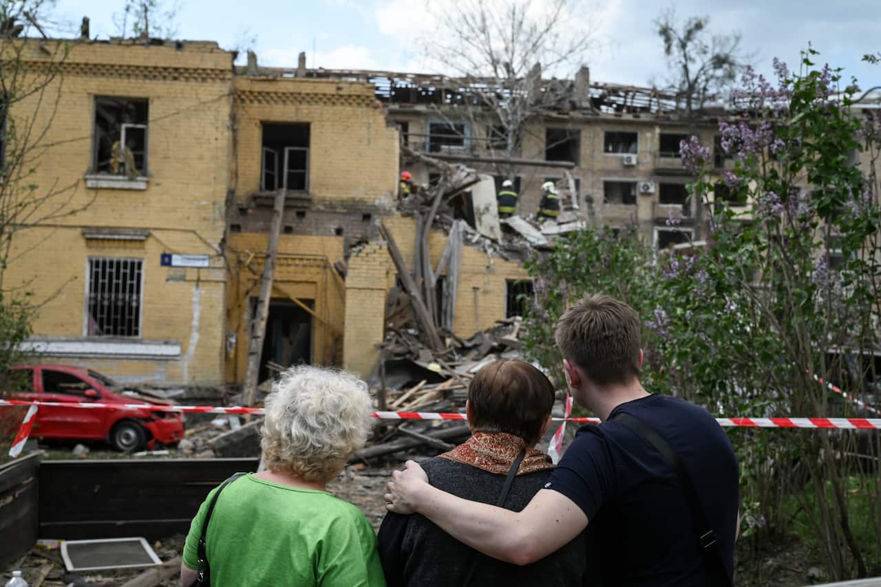 Family members embrace outside crushed building. 