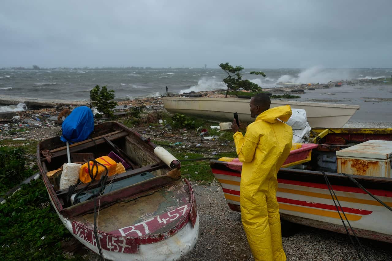 A man in a yellow raincoat stands next to boats and debris that have washed up on a shore, amid a storm.
