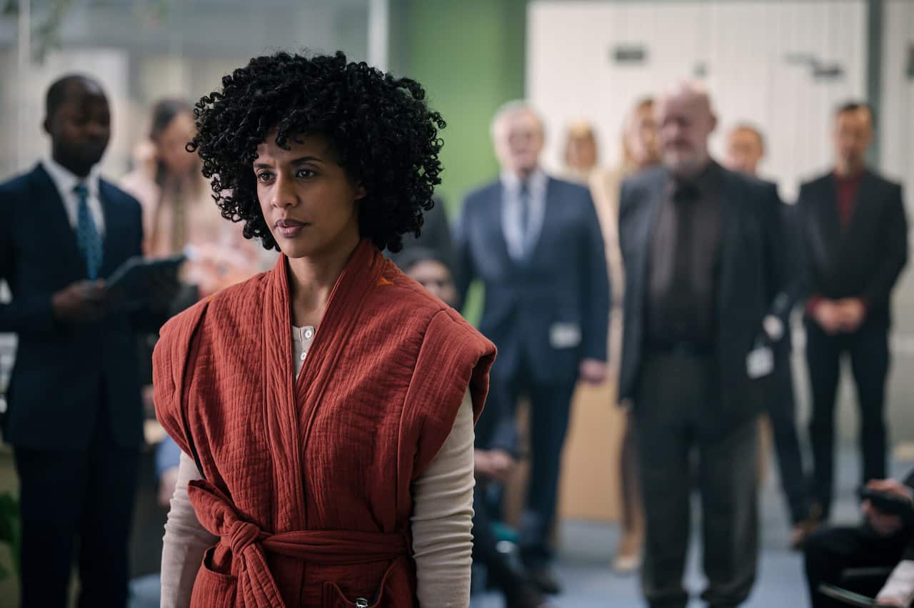 A woman in a red dress tands in front of a group of men in suits. 