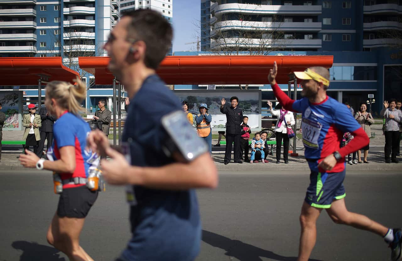 A runner wearing a blue and red top waves at people on the side of a street. Two other runners in similar clothing are ahead of him.