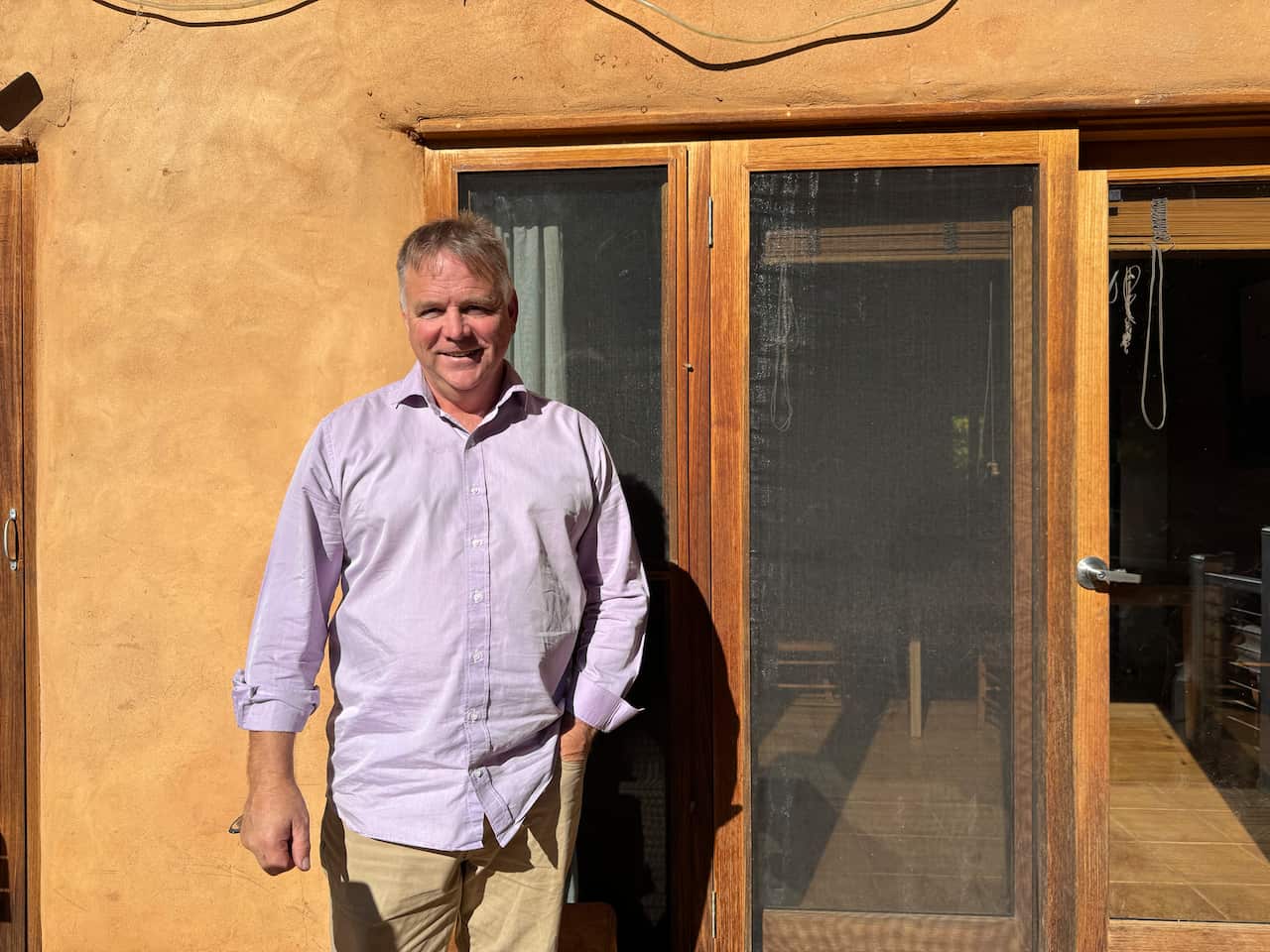 A man in a light blue shirt and fawn trousers with one hand in his pocket stands next to a straw and mud house.