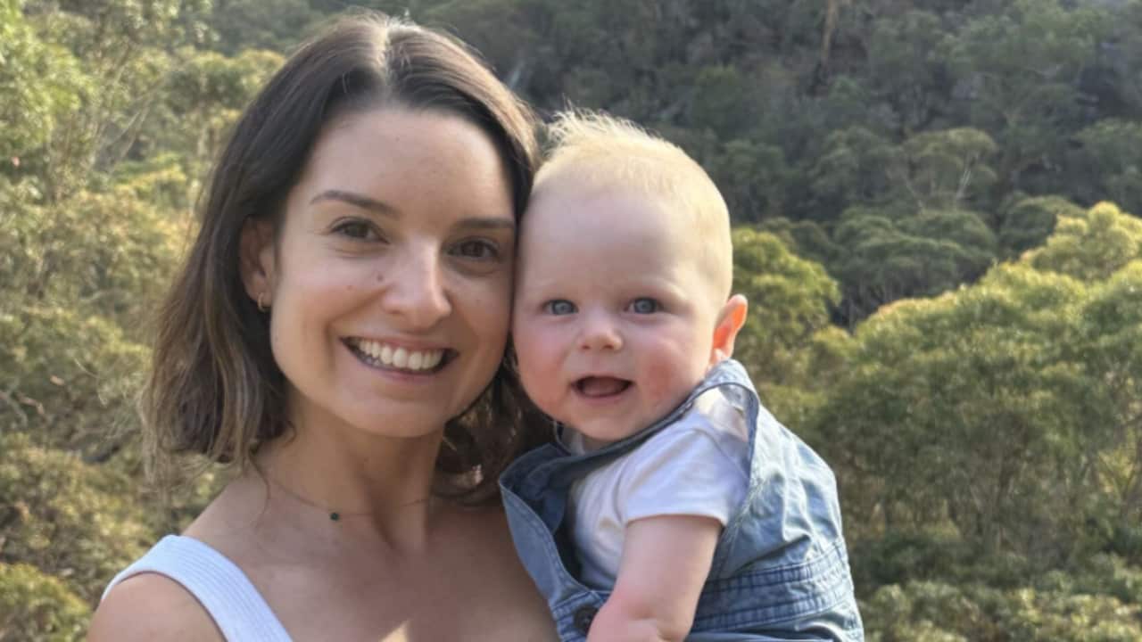 a young woman with brown hair holds a blonde baby in front of a bush landscape