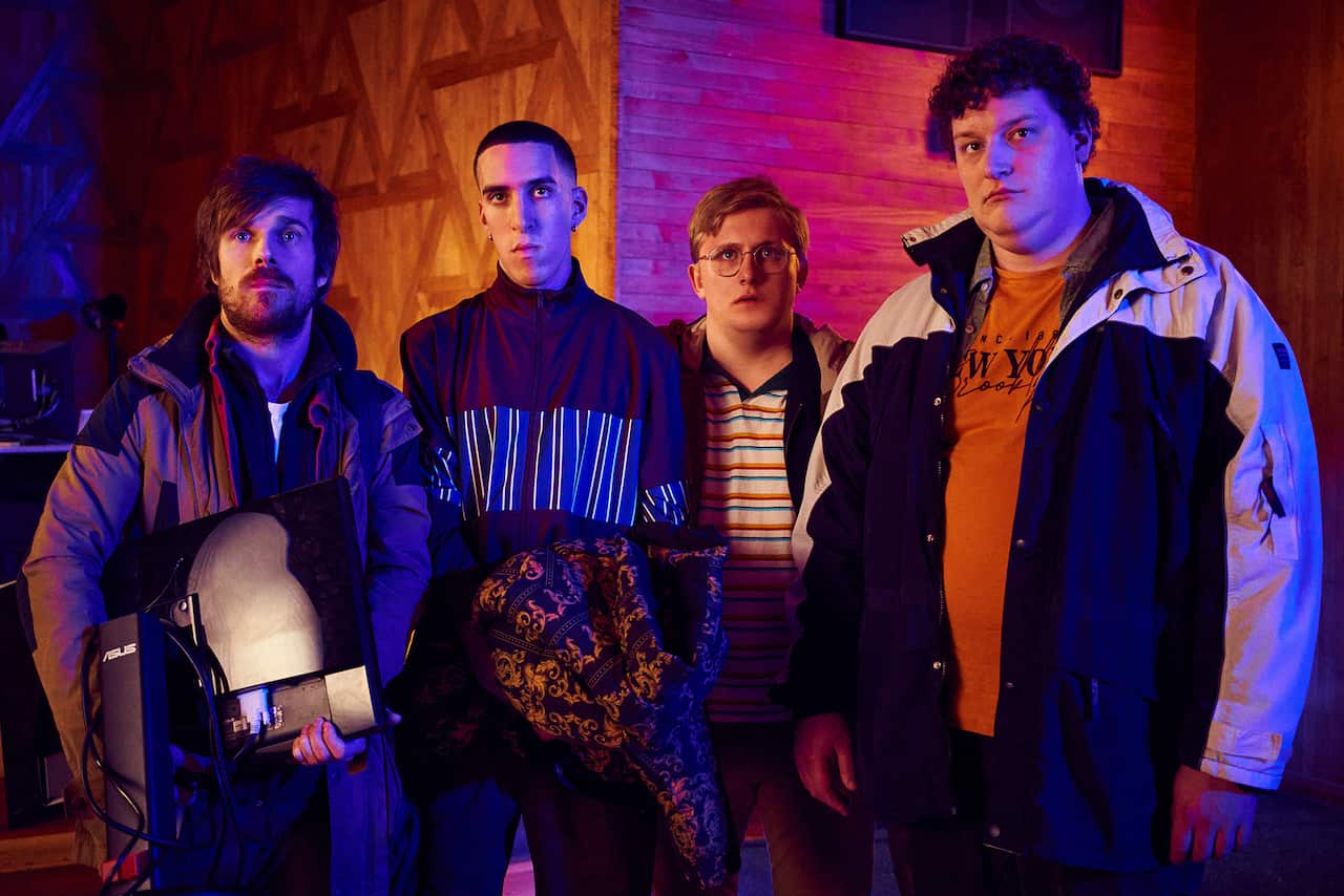 Four young men stand in a room with pink and orange lighting. One of them is holding a computer monitor.