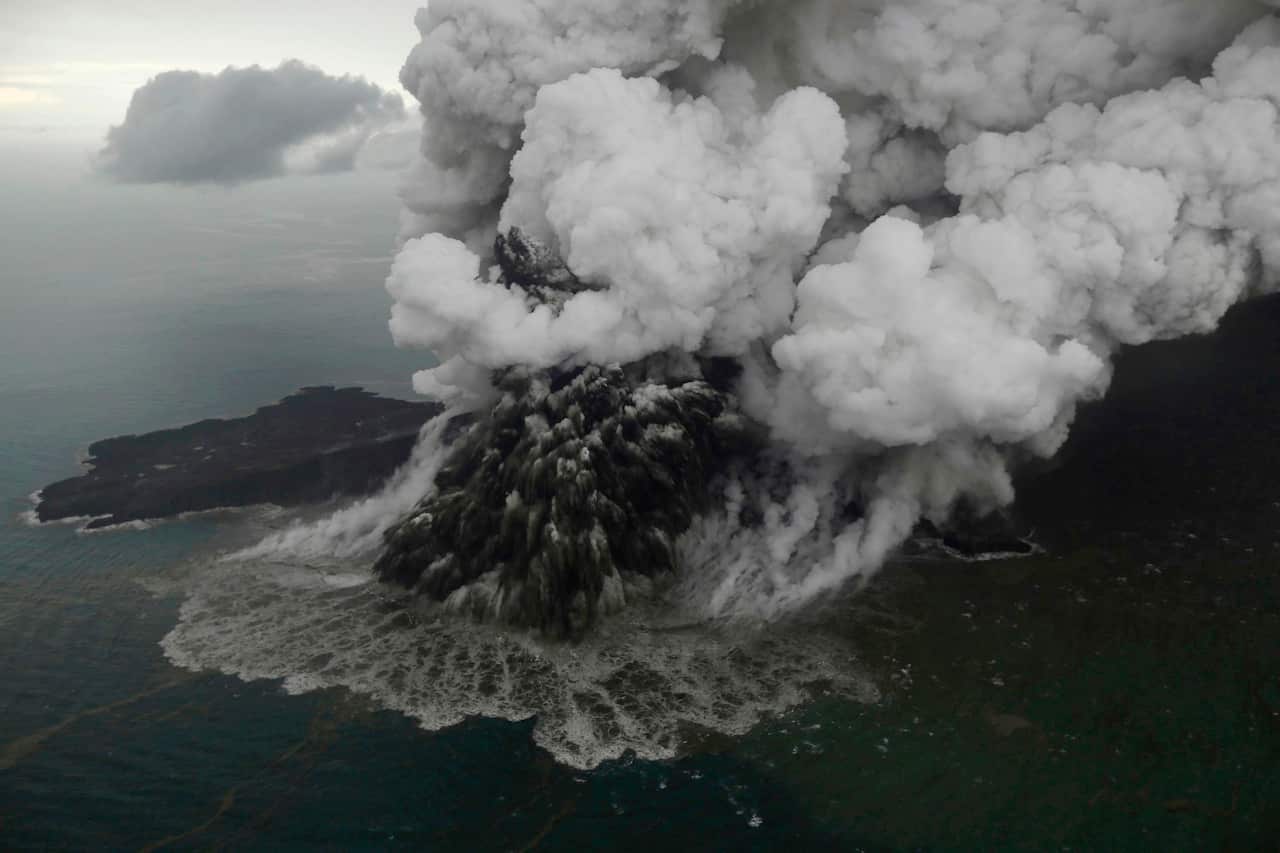 Aerial view of a volcanic eruption at sea, with dense ash and steam clouds billowing upward as dark debris and water surge around the crater.