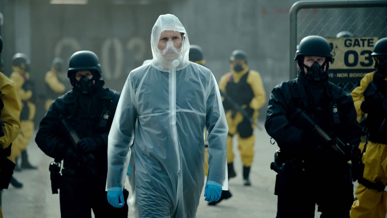 A man in a white hazard suit walks in a yard, surrounded by armed guards.