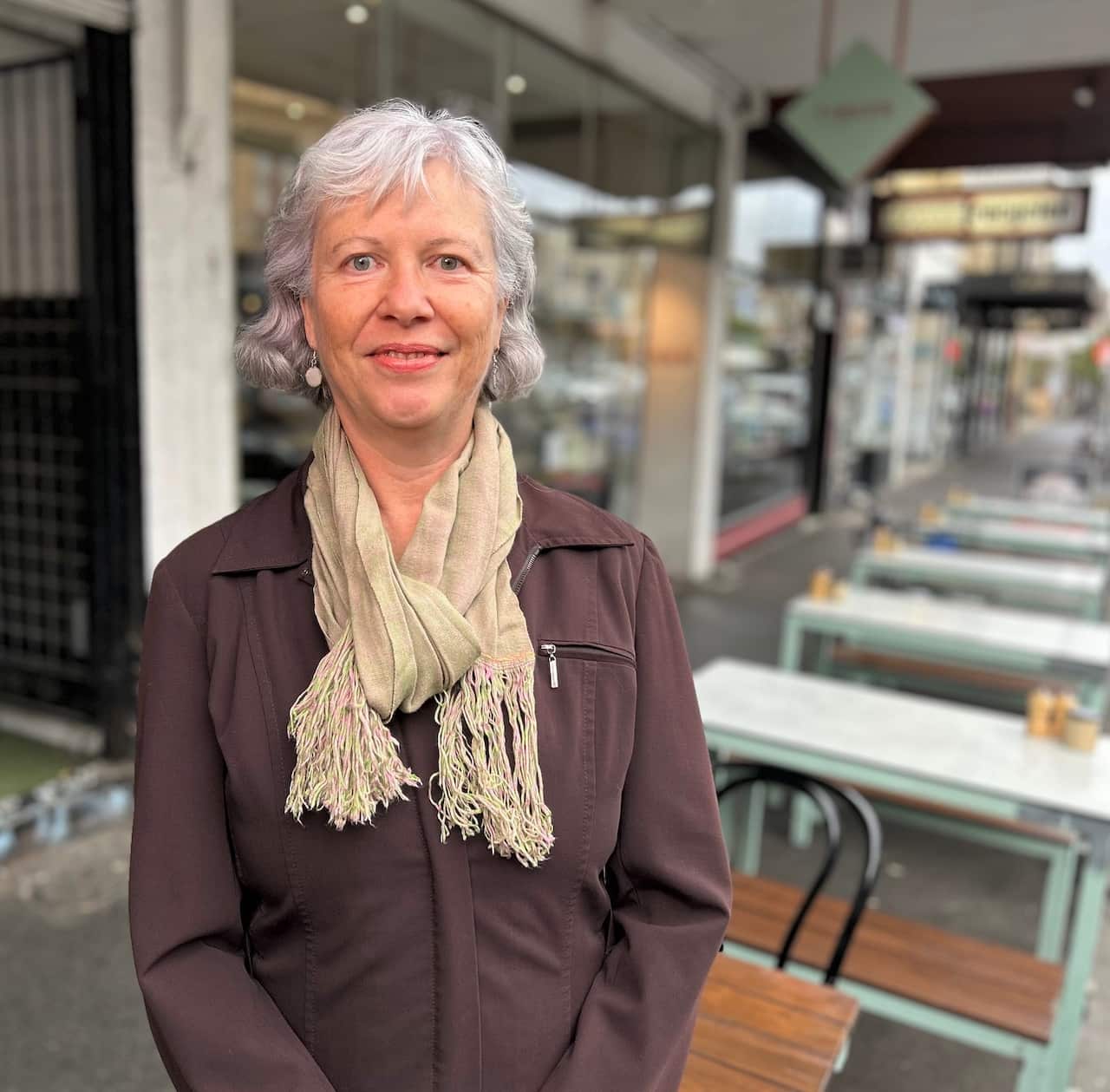 A woman in a brown jacket stands at cafe tables on a shopping street. 