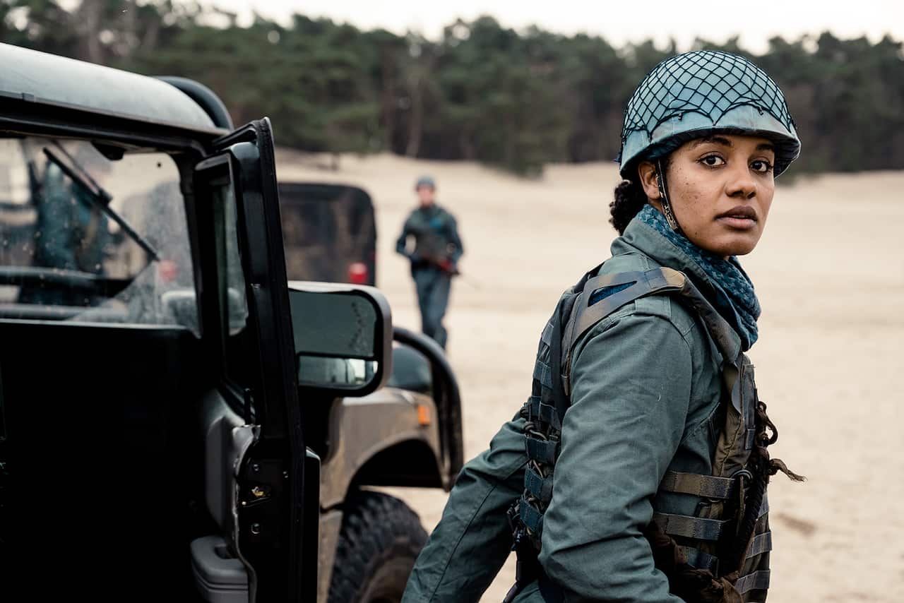 A woman in military uniform stands beside a vehicle; her body and head are turned as if she has just reacted to something. 