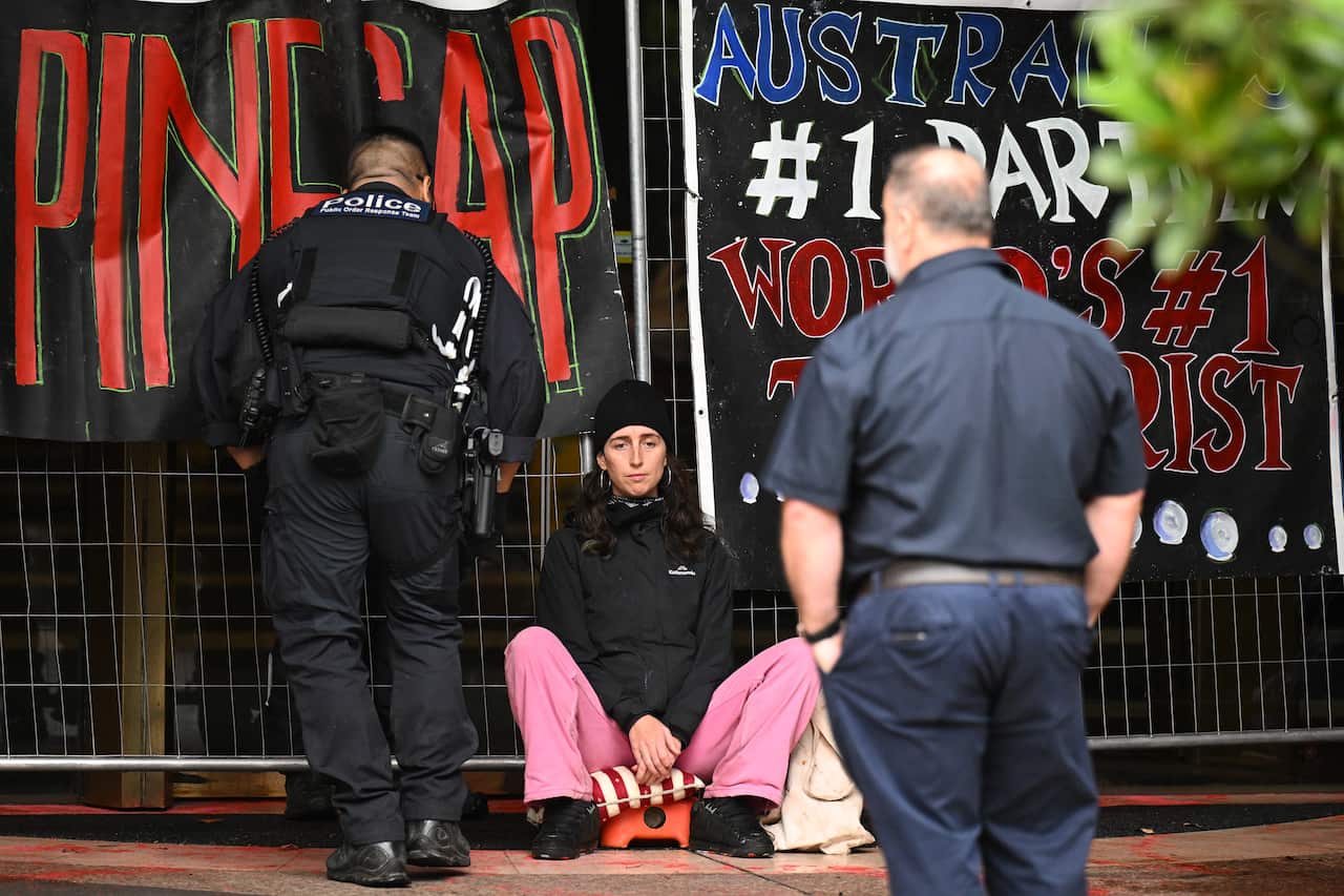 Two policemen are approaching a woman in a black jacket sitting close to a temporary fence with banners affixed.