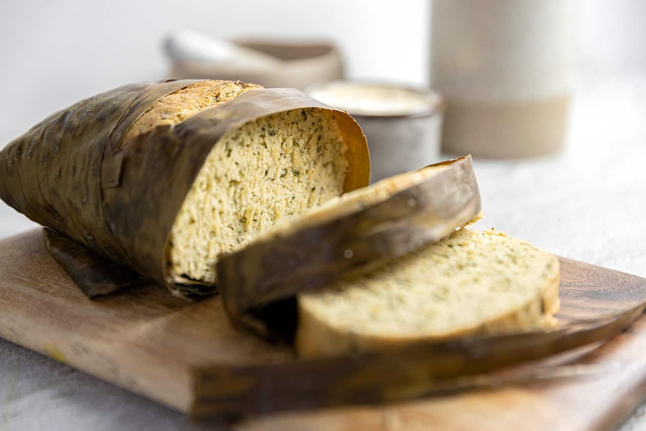 A damper, wrapped in the banan leaf it was cooked in, sits on a wooden board. Several slices have been cut, showing the green-flecked golden interior. 