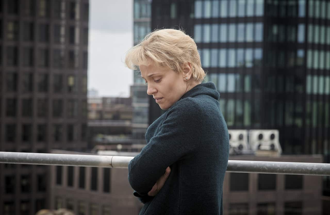 A woman with short blonde hair, wearing a blue coat, stands beside a railing, head bowed, with a several tall city buildings behind her.  