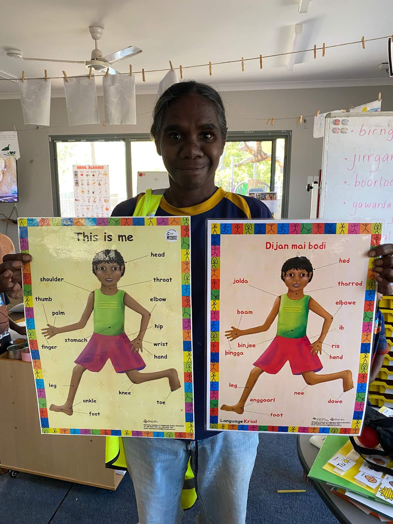 A person holding two phonics charts - one in English, one in Gija Kriol. The charts feature a picture of a child wearing a lime green singlet and red shorts with words around them indicating their main body parts.