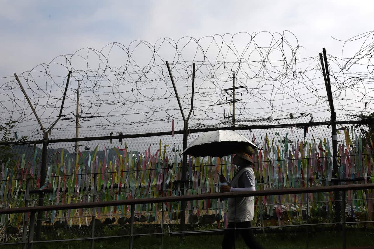 A woman with an umbrella walks past a tall barbed wire fence, which is covered in colourful prayer ribbons. 
