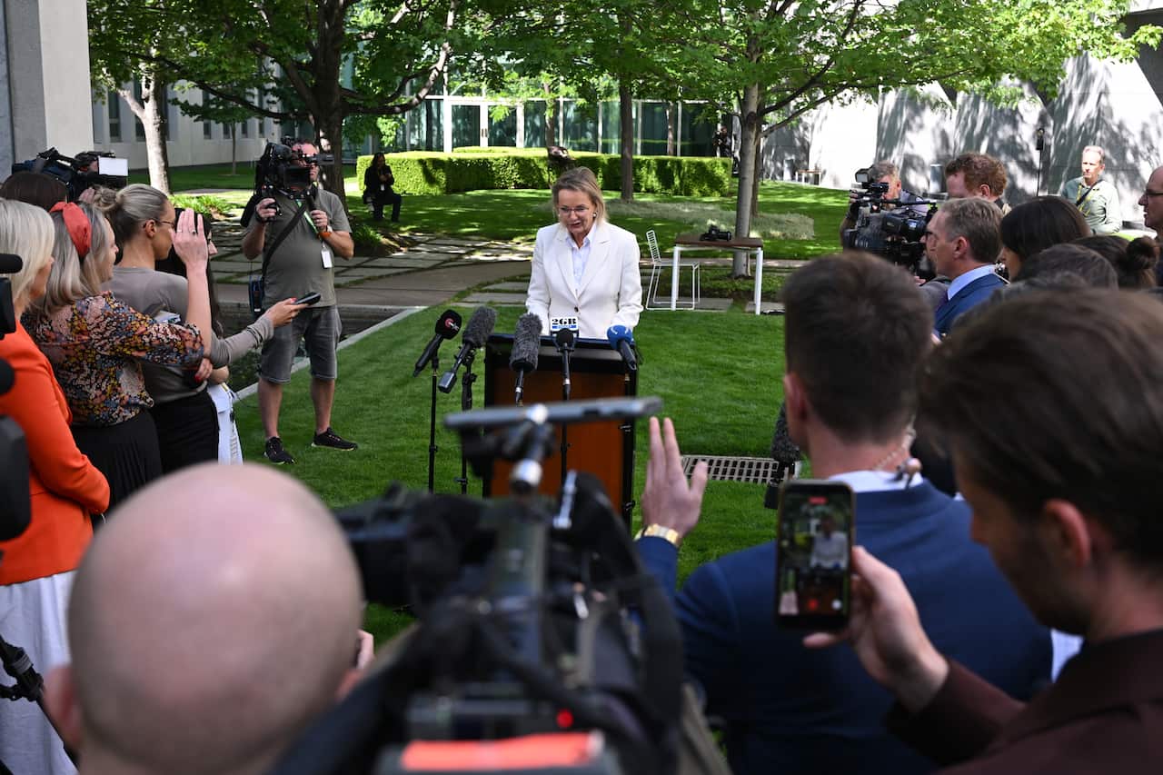 A woman dressed in a white suit speaking at press conference, surrounded by reporters and cameras