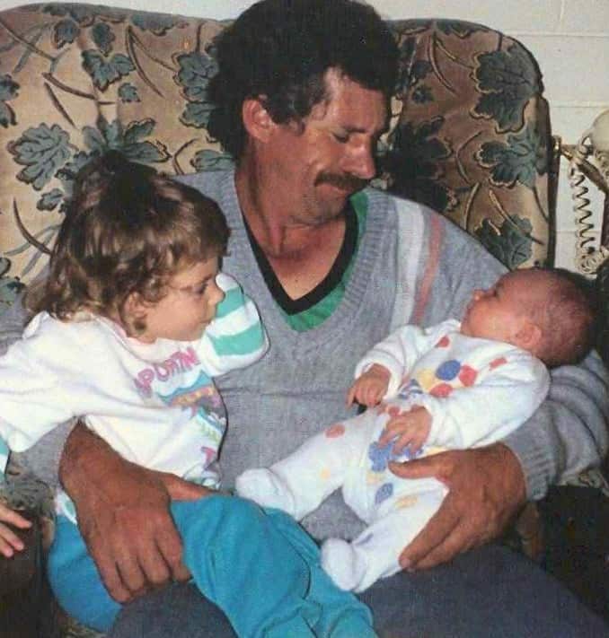 A man in a grey sweater holds a baby while a small child looks on.