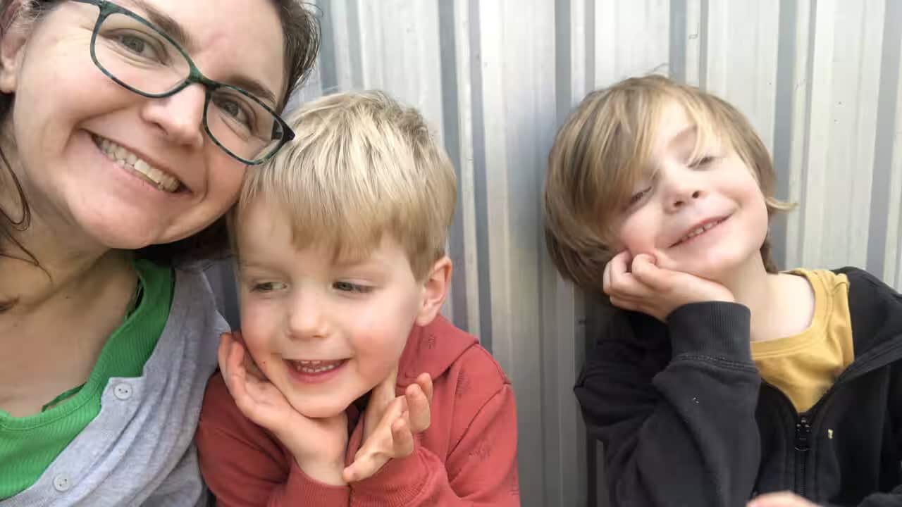 A middle-aged white woman in casual clothes is taking a selfie with her two young blonde sons, dressed in hoodies.