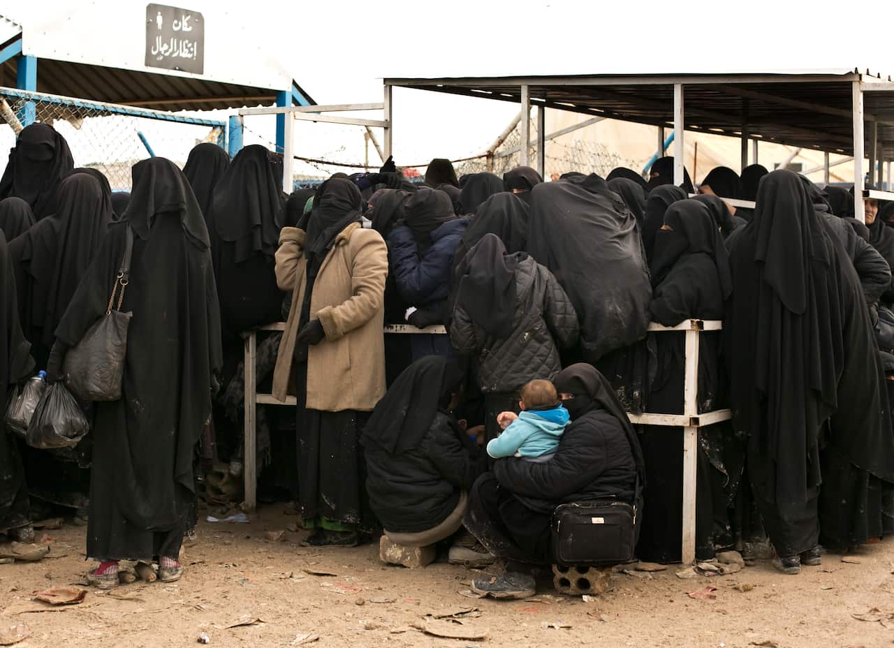 A number of women dressed in black hijabs waiting for supplies outside a Syrian camp. One of the crouched women holds a baby in her arms.