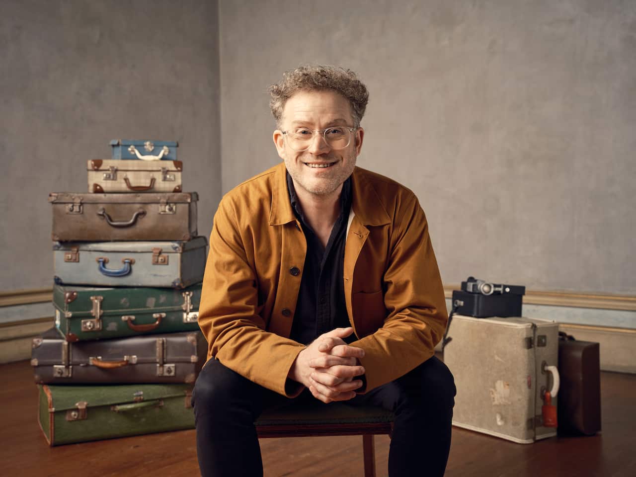 A man with glasses is sitting down and smiling. There is a stack of suitcases behind him.